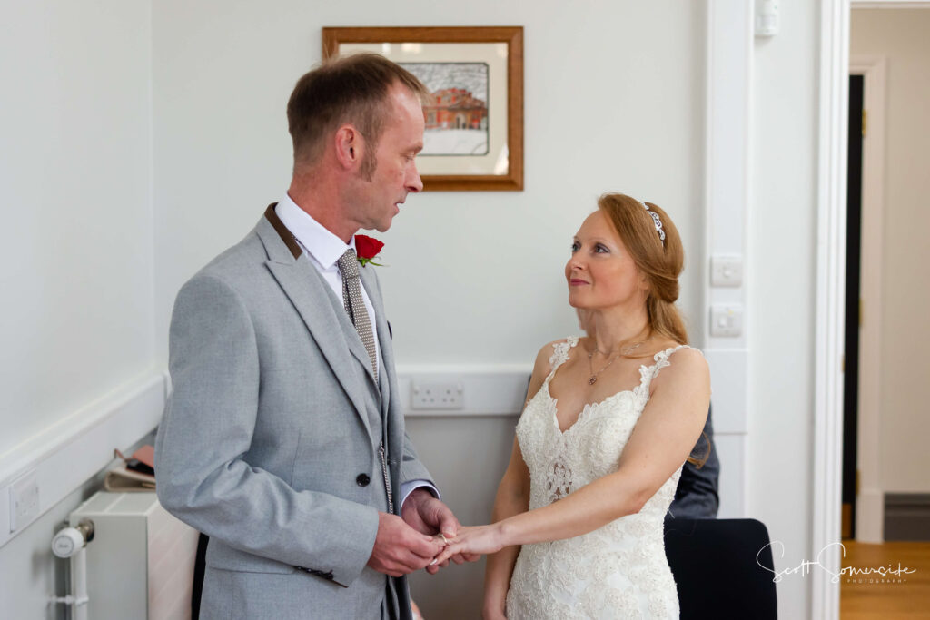 A man in a grey suit and a woman in a white wedding dress hold hands and look at each other in a brightly lit room. Copyright Somerside Photography Ltd // www.somersidephotography.com