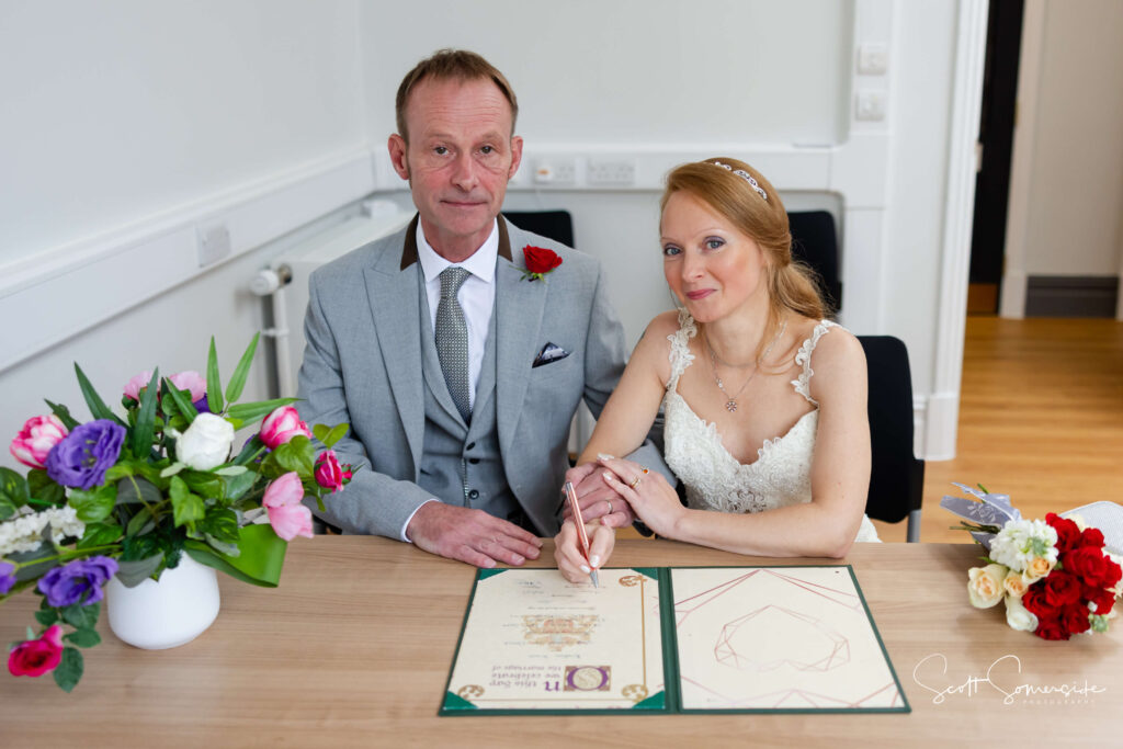 A man in a grey suit and a woman in a white dress sit at a table, signing a document with flowers nearby. Copyright Somerside Photography Ltd // www.somersidephotography.com
