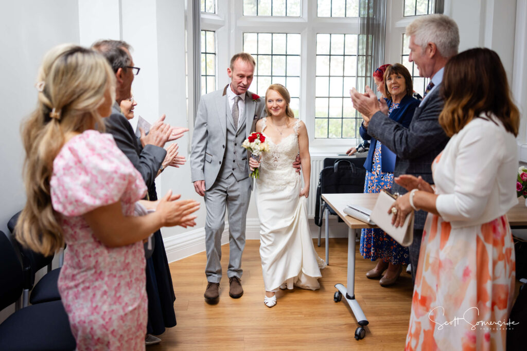 A bride and groom walk together down an aisle in a bright room as guests standing on either side applaud them. Copyright Somerside Photography Ltd // www.somersidephotography.com