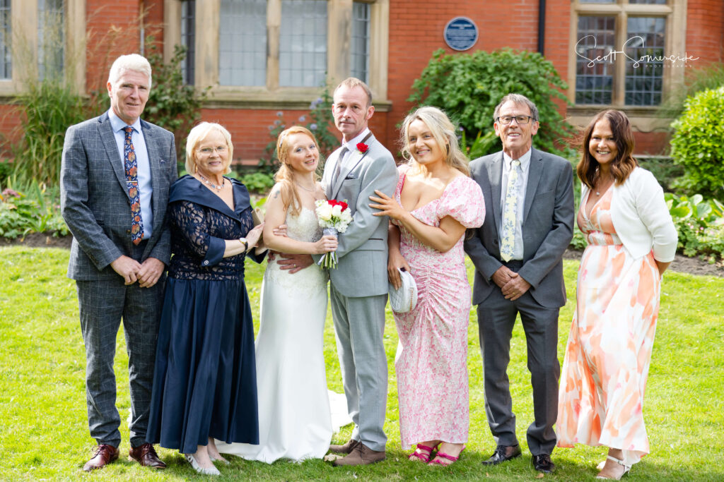 A bride and groom stand together holding a bouquet, surrounded by four adults and one woman in a floral dress, all posing for a group photo outdoors in front of a brick building. Copyright Somerside Photography Ltd // www.somersidephotography.com