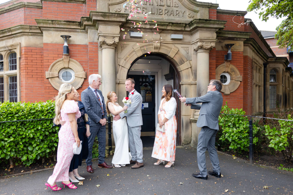 A small wedding group stands outside a historic brick building as a man tosses flower petals over a newly-married couple at the entrance. Copyright Somerside Photography Ltd // www.somersidephotography.com