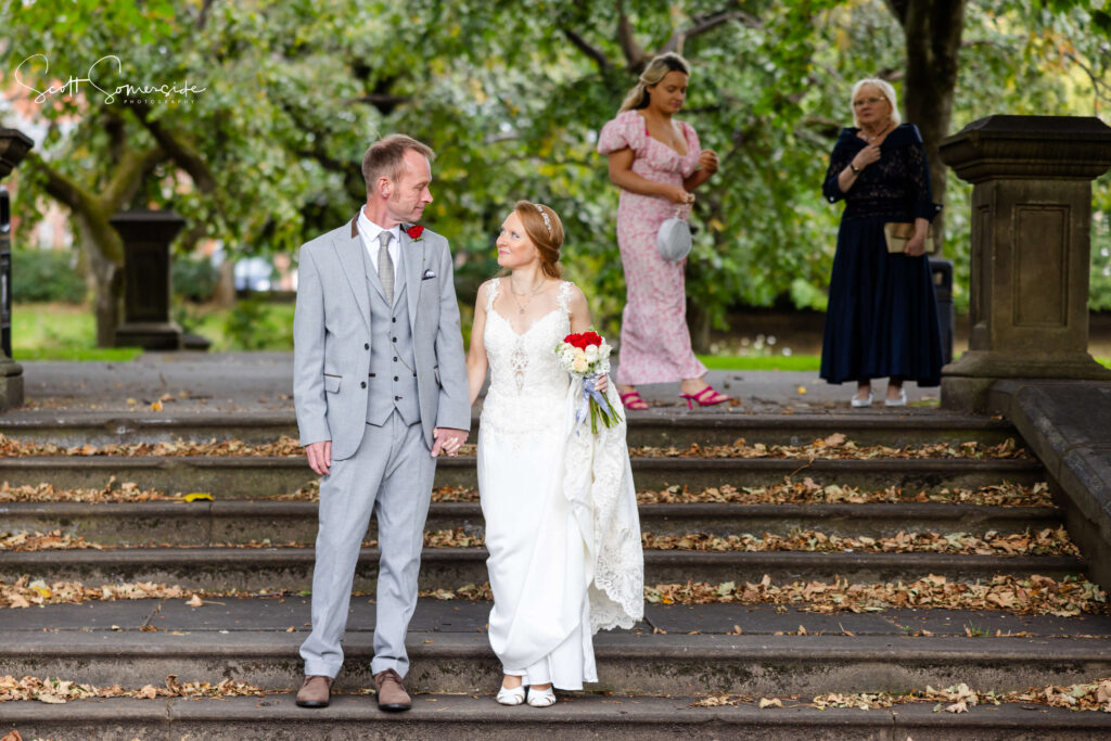 A bride and groom walk down stone steps outdoors, holding hands. Two women stand in the background, one holding a mobile. Trees with green leaves frame the scene. Copyright Somerside Photography Ltd // www.somersidephotography.com