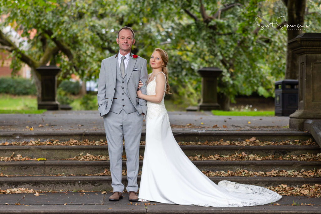 A bride in a white gown stands next to a groom in a grey suit on stone steps outdoors, with green trees in the background. Copyright Somerside Photography Ltd // www.somersidephotography.com