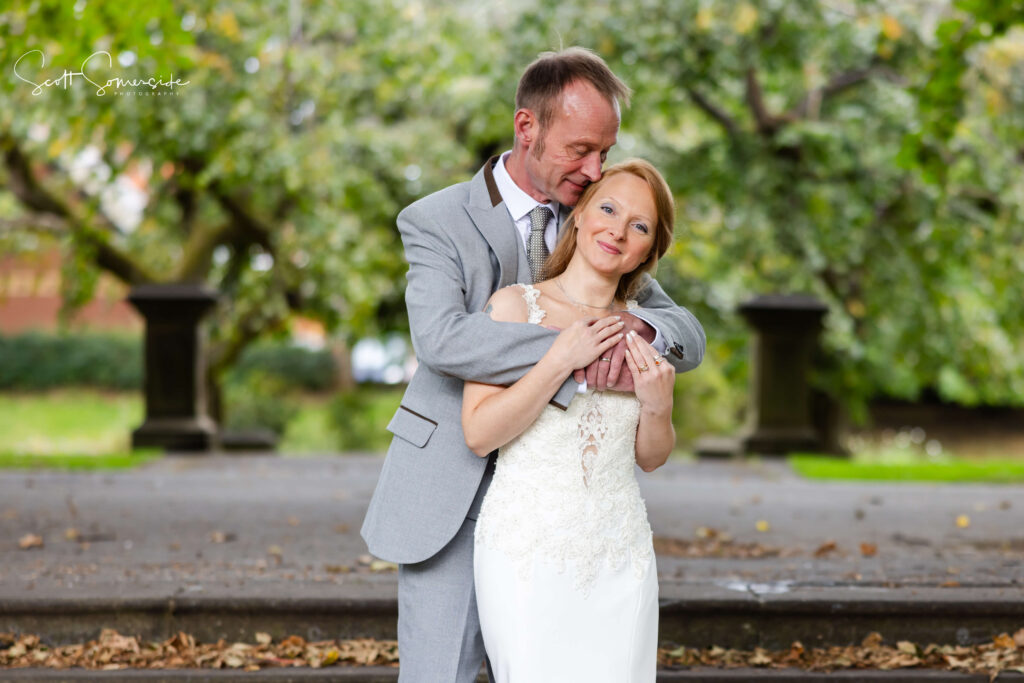 A man in a grey suit embraces a woman in a white wedding dress whilst standing outdoors in a park with trees in the background. Copyright Somerside Photography Ltd // www.somersidephotography.com