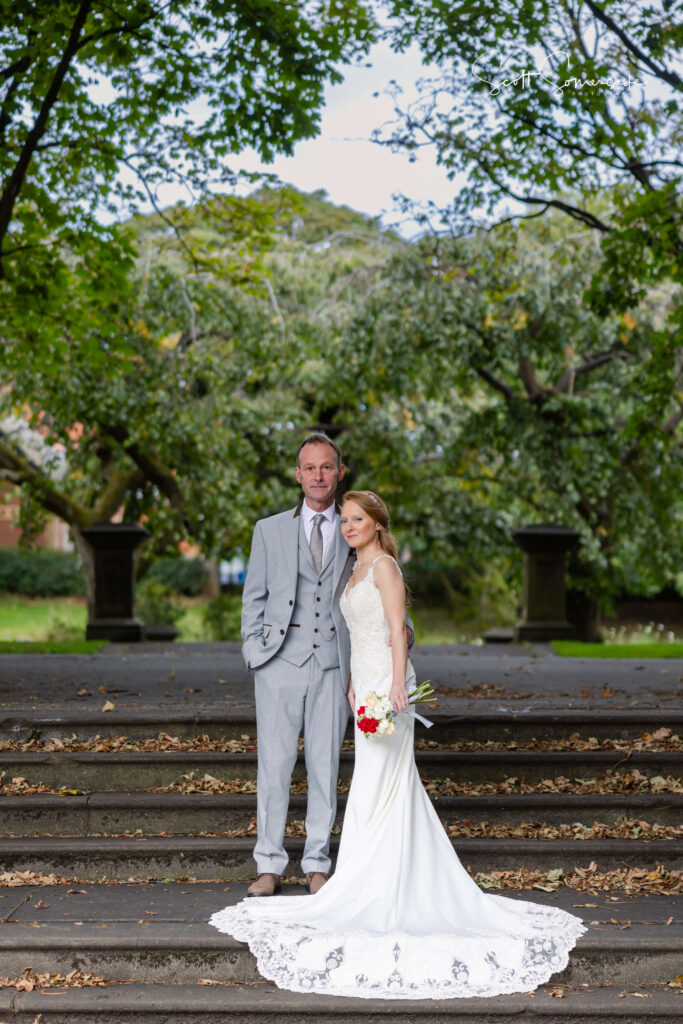 A man in a grey suit and a woman in a white wedding dress stand together on stone steps in a park, surrounded by green trees. Copyright Somerside Photography Ltd // www.somersidephotography.com