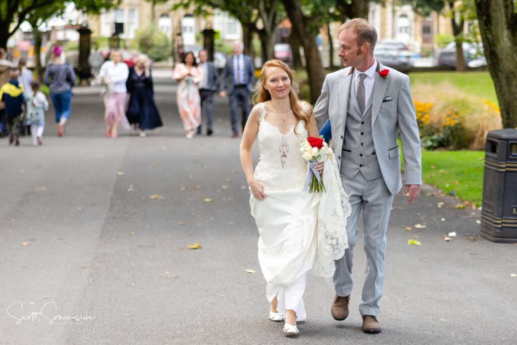 A bride in a white dress and a groom in a grey suit walk together in a park, followed by wedding guests in the background. Copyright Somerside Photography Ltd // www.somersidephotography.com