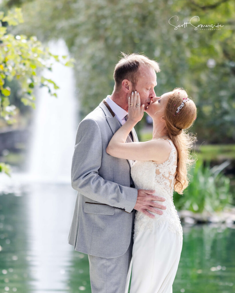 A bride and groom kiss by a pond with a waterfall in the background, surrounded by greenery. Copyright Somerside Photography Ltd // www.somersidephotography.com