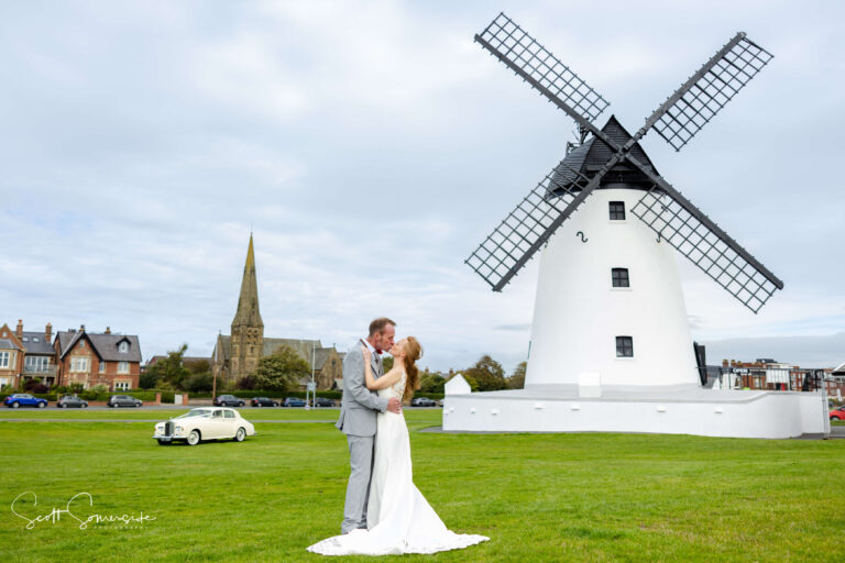 A bride and groom embrace on grass with a white windmill, church, classic car, and houses in the background under a cloudy sky. Copyright Somerside Photography Ltd // www.somersidephotography.com
