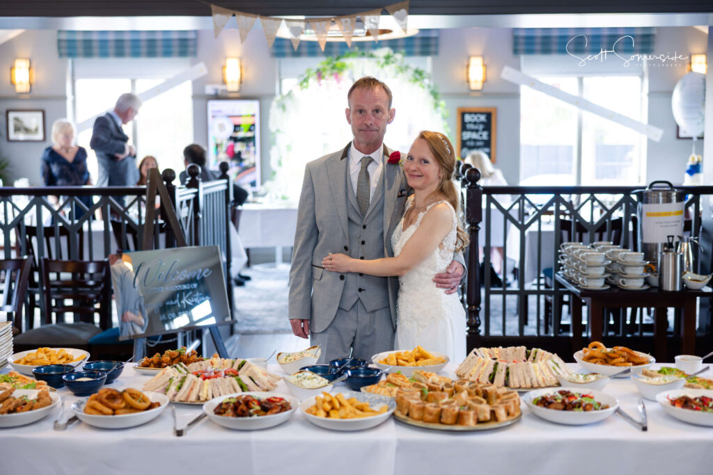 A bride and groom stand together in front of a large table of assorted party food at a wedding reception in a decorated indoor venue. Copyright Somerside Photography Ltd // www.somersidephotography.com