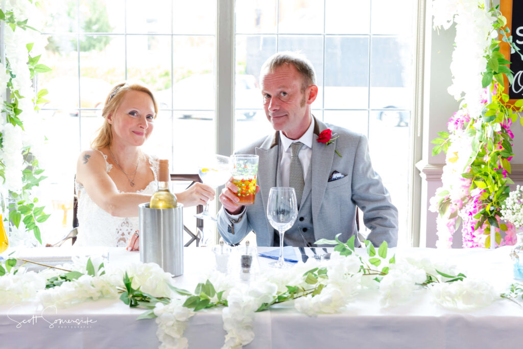 A bride and groom sit at a decorated table, raising glasses for a toast, with white floral arrangements and a wine bottle in an ice bucket. Copyright Somerside Photography Ltd // www.somersidephotography.com