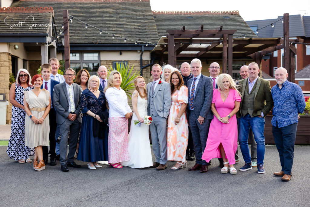 A wedding group photo outside, with the bride and groom in the centre surrounded by family and friends, all dressed in formal and semi-formal attire. Copyright Somerside Photography Ltd // www.somersidephotography.com