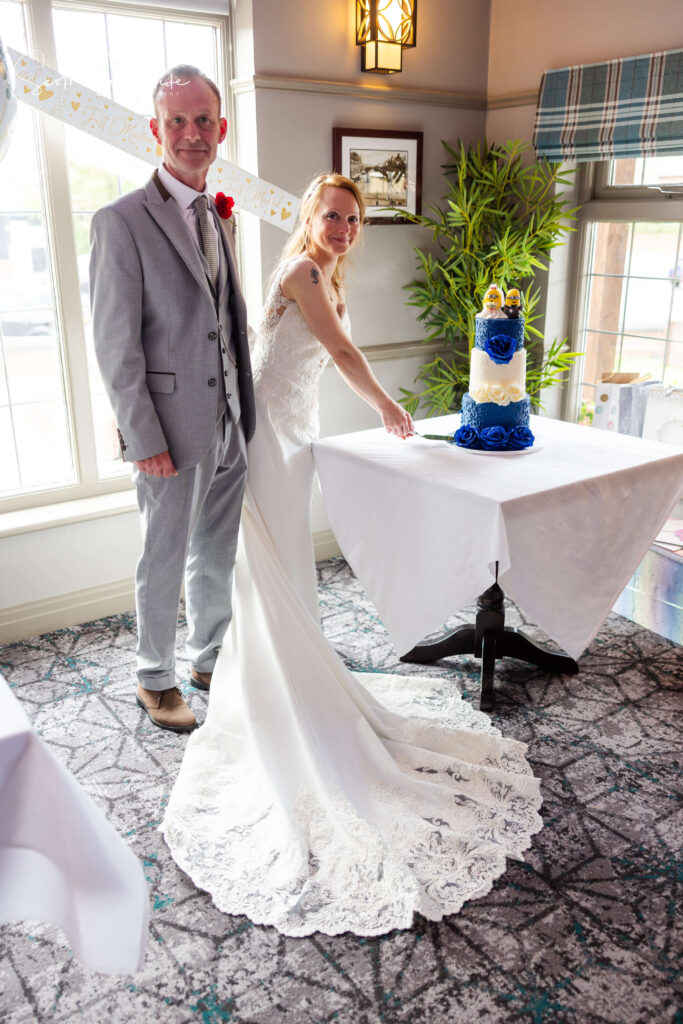 A bride and groom stand together indoors by a table, as the bride prepares to cut a decorated wedding cake. Copyright Somerside Photography Ltd // www.somersidephotography.com