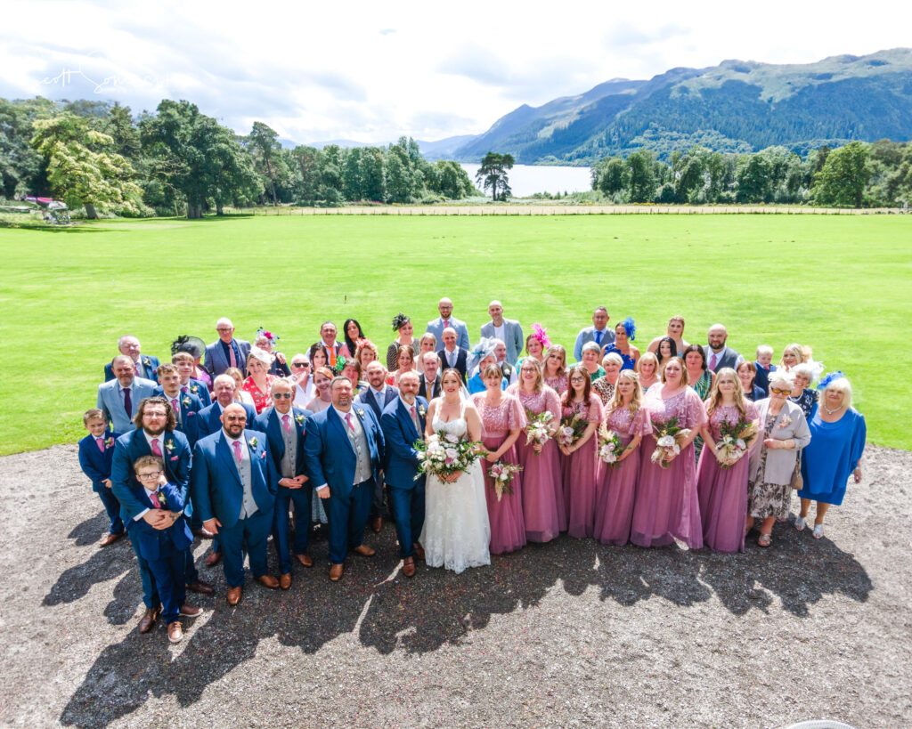 Large wedding party poses outdoors, with bridesmaids in mauve dresses and groomsmen in blue suits, on a gravel area with green lawn, trees, and hills in the background. Copyright Somerside Photography Ltd // www.somersidephotography.com