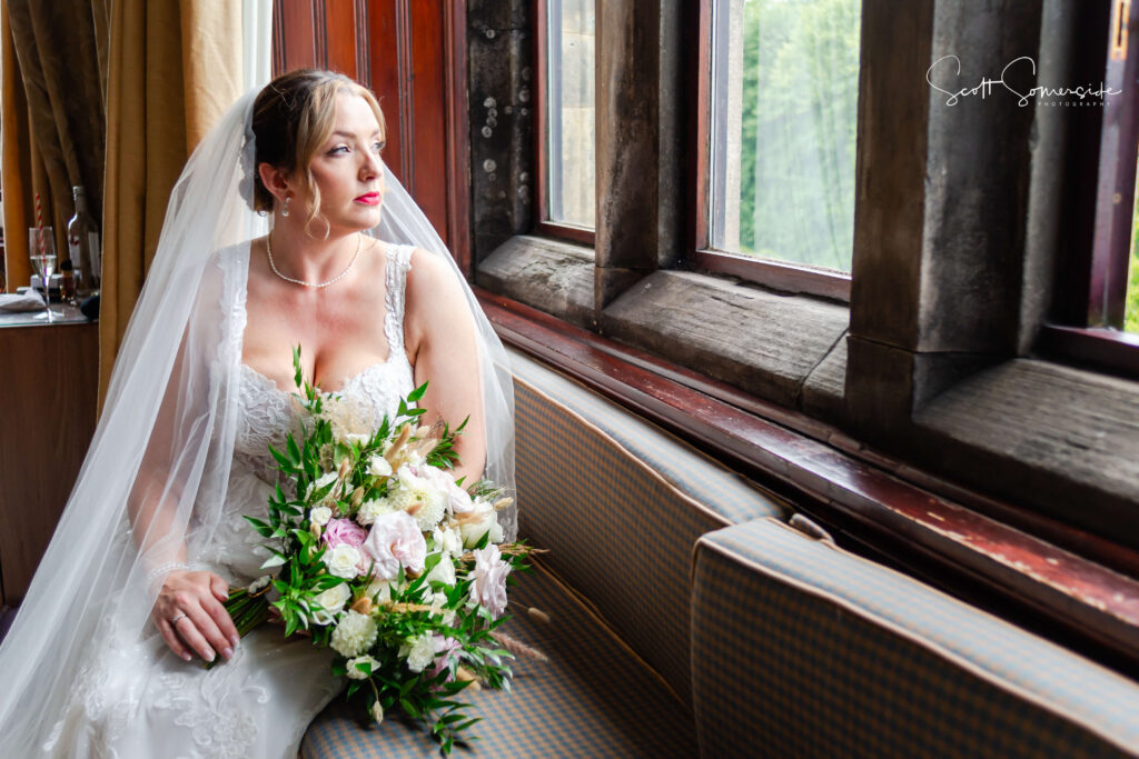 A bride in a white dress and veil sits on a bench by a window, holding a bouquet of flowers and looking outside. Copyright Somerside Photography Ltd // www.somersidephotography.com