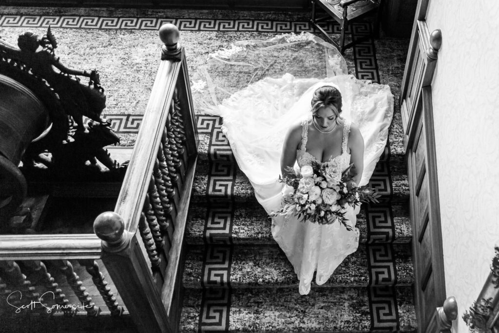 A bride in a wedding dress holding a bouquet walks down a carpeted staircase, viewed from above in black and white. Copyright Somerside Photography Ltd // www.somersidephotography.com