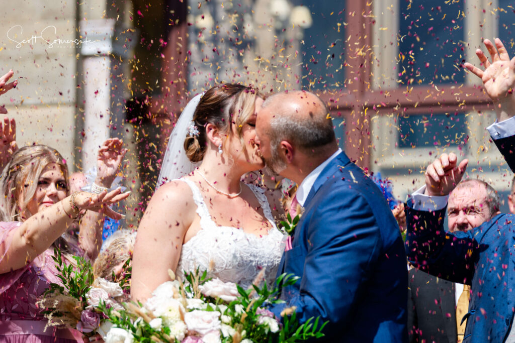 A bride and groom kiss outdoors amid falling confetti, surrounded by cheering guests. The bride holds a bouquet and wears a white dress; the groom is in a blue suit. Copyright Somerside Photography Ltd // www.somersidephotography.com