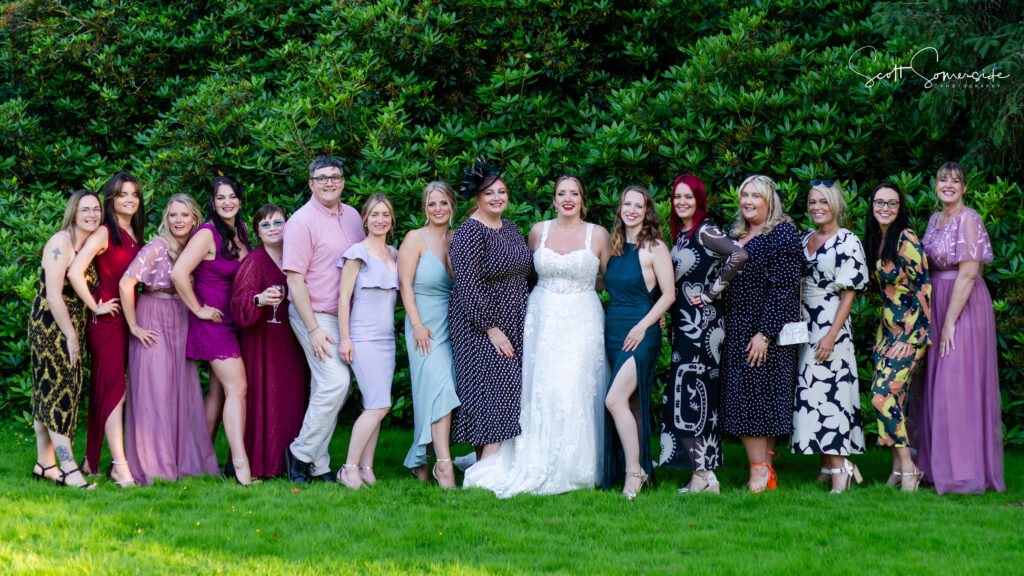 A group of people, including a bride in a white dress, pose together outdoors on grass in front of green foliage. Copyright Somerside Photography Ltd // www.somersidephotography.com