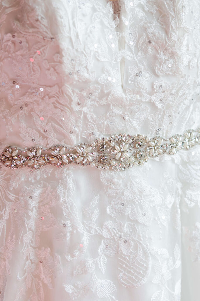 Close-up of a white lace wedding dress with sequin details, featuring a jewelled belt at the waist. Copyright Somerside Photography Ltd // www.somersidephotography.com