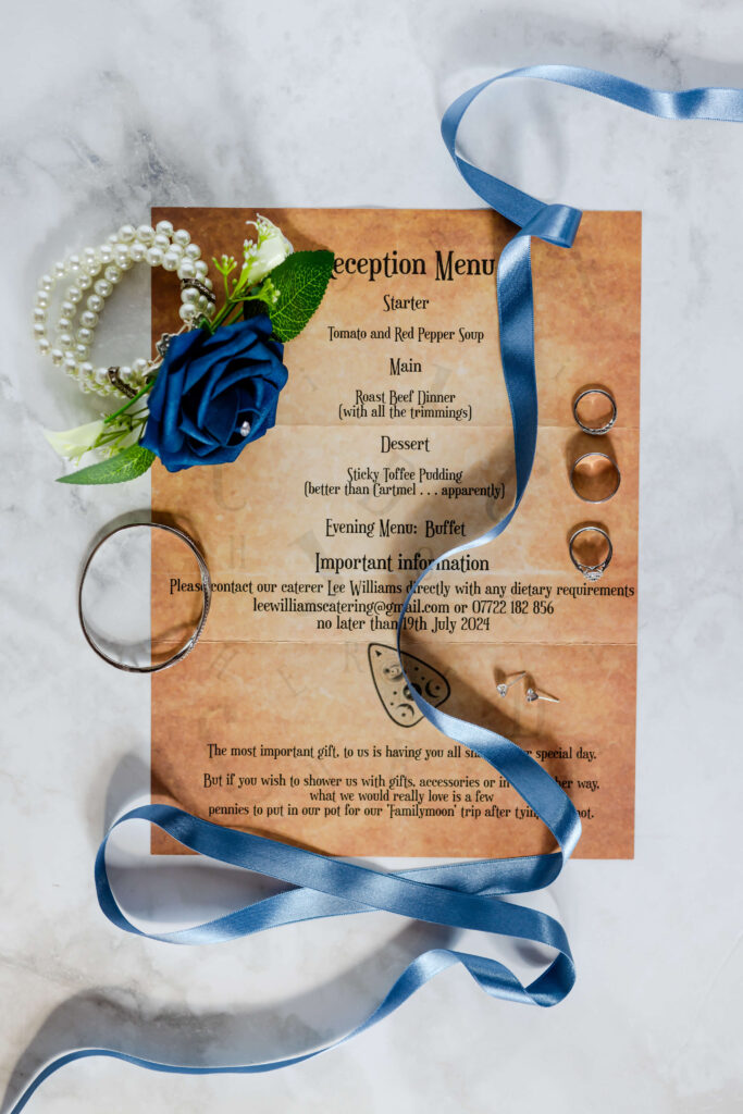 A wedding reception menu on brown paper with gold rings, a blue rose corsage, pearl bracelet, and blue ribbon arranged on a light surface. Copyright Somerside Photography Ltd // www.somersidephotography.com