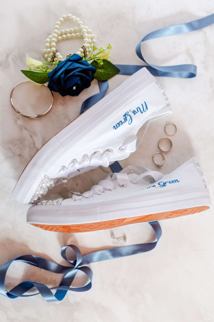 Two white trainers with "Mrs Green" embroidered in blue, surrounded by rings, a bracelet, a blue rose, pearl decor, and blue ribbon on a marble surface. Copyright Somerside Photography Ltd // www.somersidephotography.com