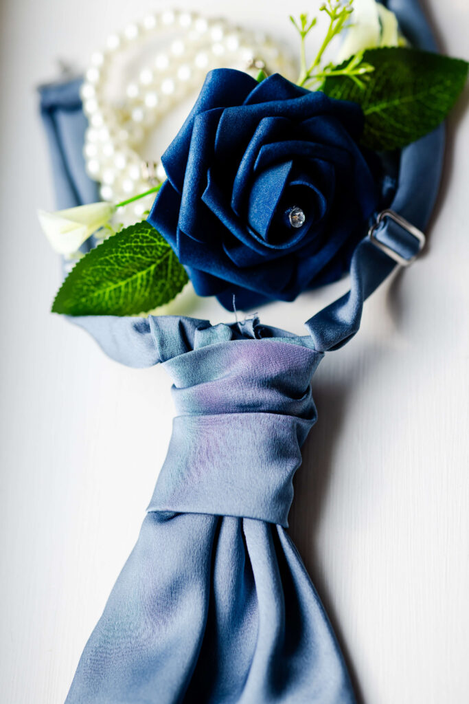 A blue necktie, an artificial blue rose with green leaves, and a string of white pearls are arranged on a light surface. Copyright Somerside Photography Ltd // www.somersidephotography.com