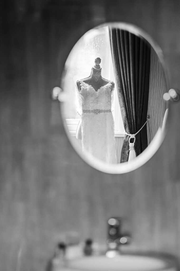 A white lace wedding dress on a mannequin is reflected in an oval mirror, with a window and curtain visible in the background. Copyright Somerside Photography Ltd // www.somersidephotography.com