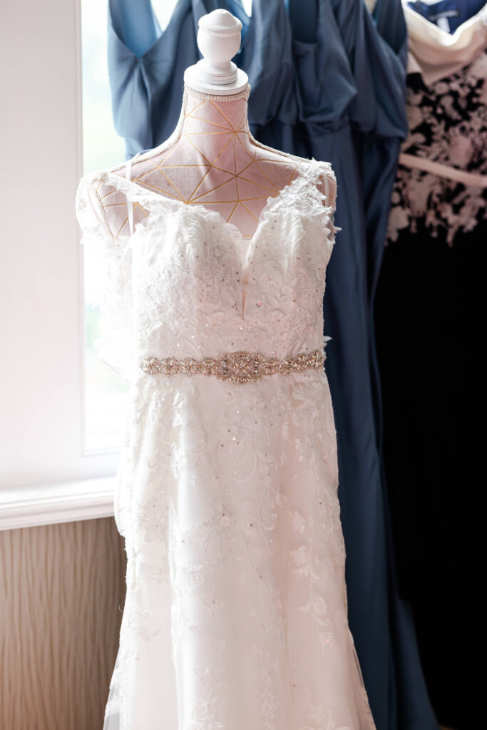 A white lace wedding dress with a jewelled belt displayed on a mannequin, with blue and black dresses hanging in the background. Copyright Somerside Photography Ltd // www.somersidephotography.com