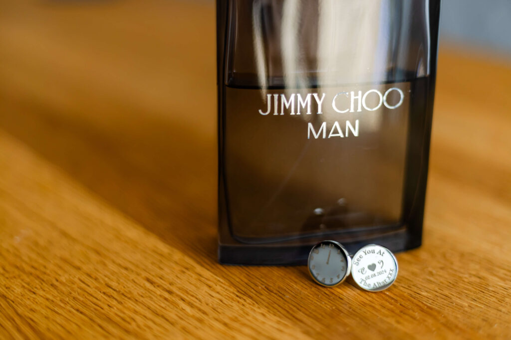 A bottle of Jimmy Choo Man aftershave stands on a wooden surface next to a small round cufflink engraved with "I love you all the time" and a small clock icon. Copyright Somerside Photography Ltd // www.somersidephotography.com