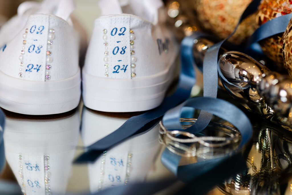 A pair of white trainers with rhinestones and the date "02 08 24" written on the heels, next to blue ribbon, wedding rings, and decorative items on a reflective surface. Copyright Somerside Photography Ltd // www.somersidephotography.com