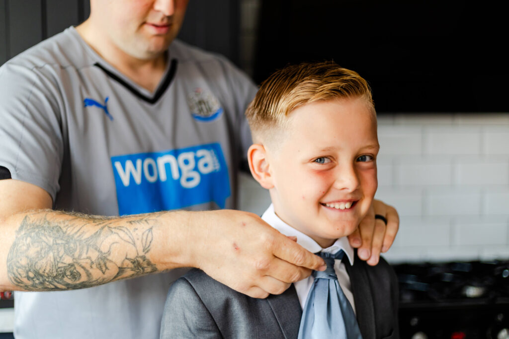 An adult adjusts a young boy’s tie while he smiles, wearing a suit. The adult has tattoos and wears a Newcastle United football shirt. Copyright Somerside Photography Ltd // www.somersidephotography.com