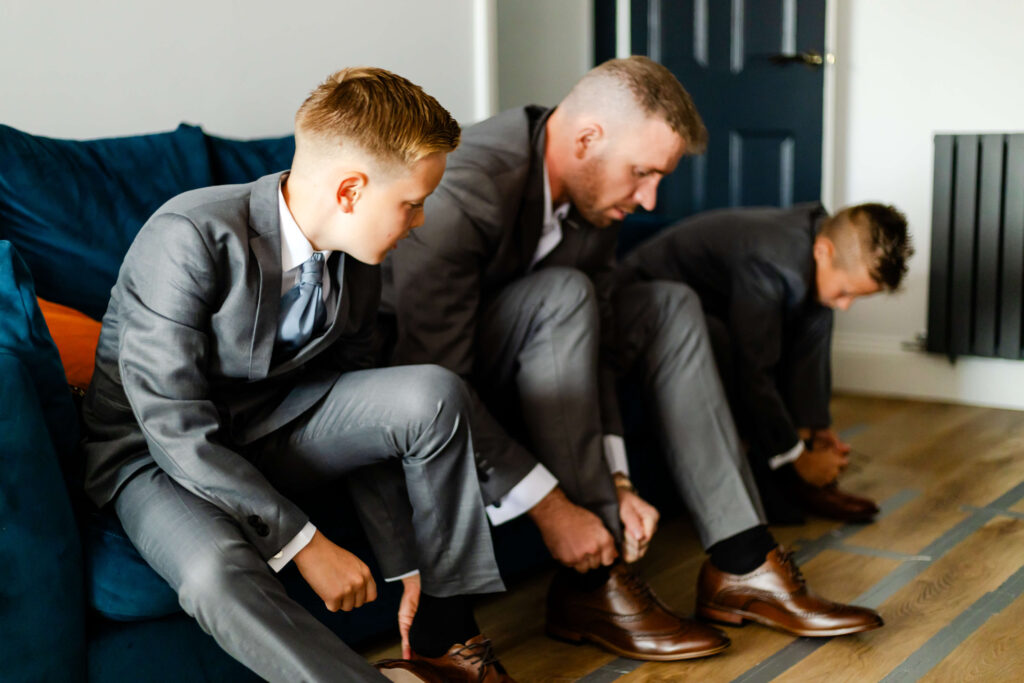 Three people in grey suits sit indoors, bending down to tie their brown dress shoes; two boys and one adult are visible. Copyright Somerside Photography Ltd // www.somersidephotography.com