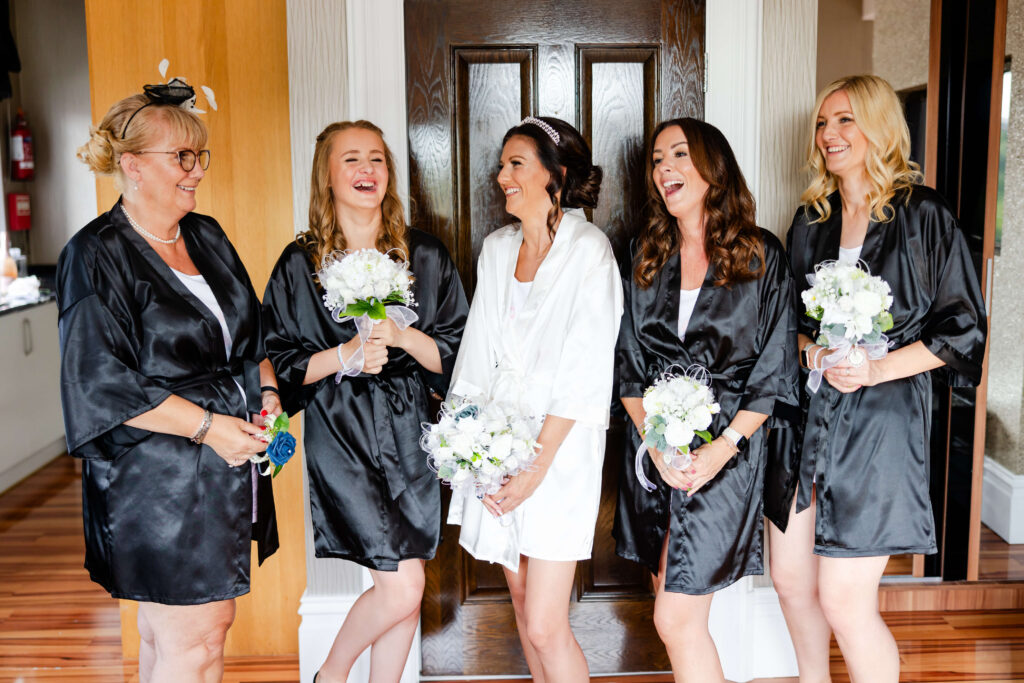 Five women in robes, four in black and one in white, stand in front of a dark wooden door, smiling and holding bouquets. Copyright Somerside Photography Ltd // www.somersidephotography.com