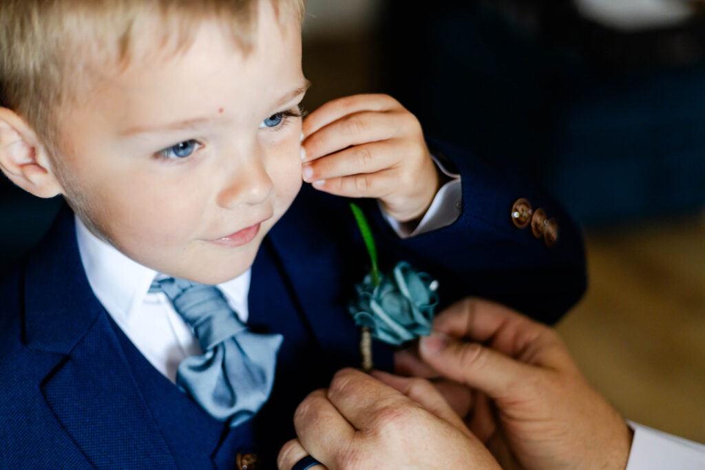 Young boy dressed in a navy blue suit with a bow tie, having a buttonhole pinned to his jacket by an adult. Copyright Somerside Photography Ltd // www.somersidephotography.com