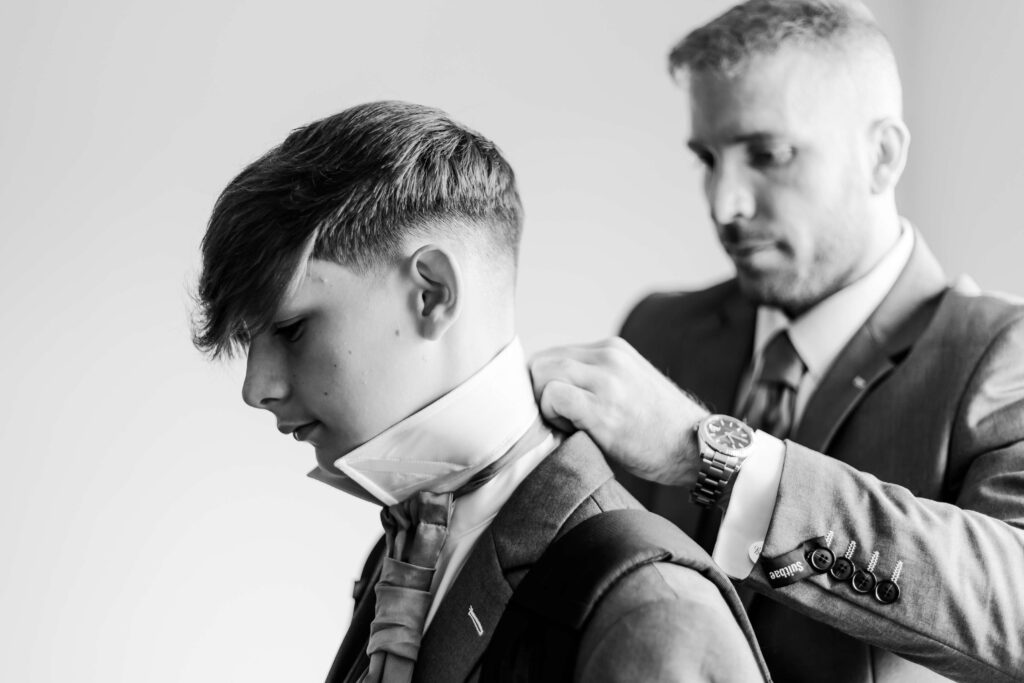 A man in a suit helps a younger person adjust a tie and collar, both wearing formal attire in a black and white photo. Copyright Somerside Photography Ltd // www.somersidephotography.com