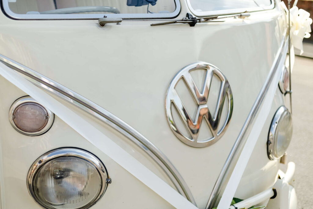 Close-up of the front of a cream-coloured vintage Volkswagen van decorated with white ribbons, showing headlamps and the VW logo. Copyright Somerside Photography Ltd // www.somersidephotography.com