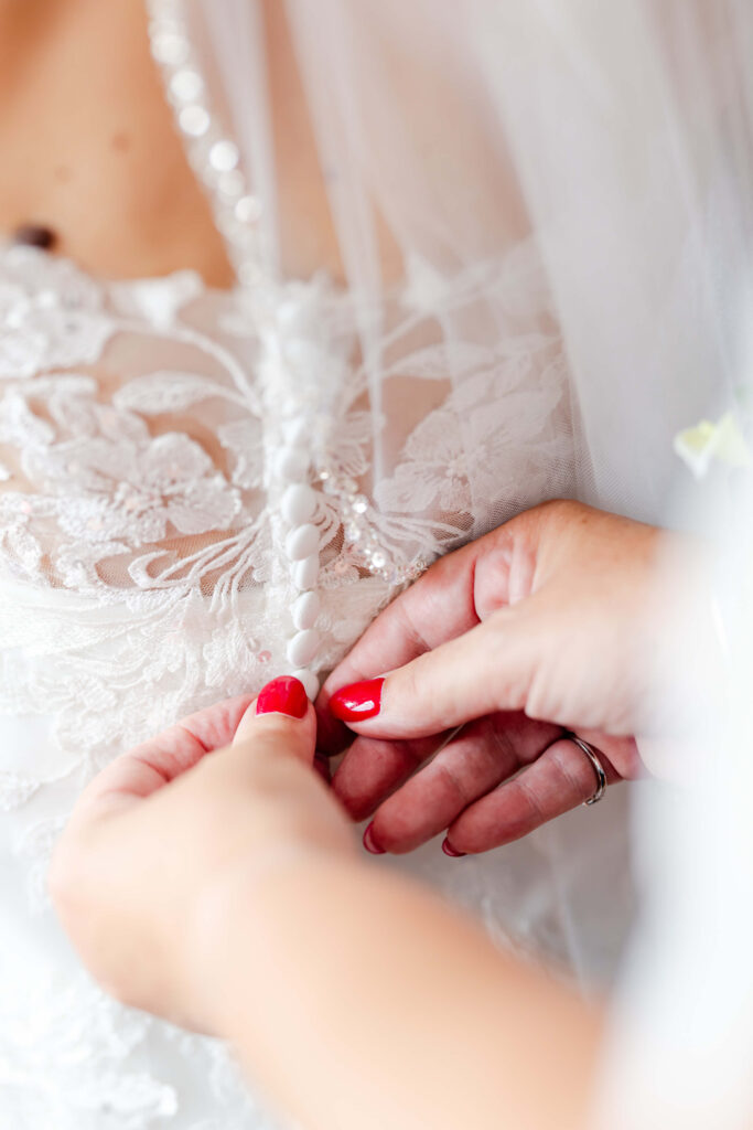 Close-up of hands with red nail varnish fastening buttons on the back of a white lace wedding dress. Copyright Somerside Photography Ltd // www.somersidephotography.com