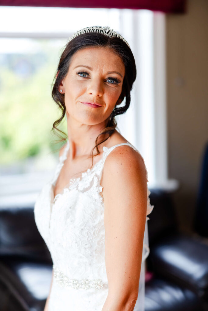 Woman in a white lace wedding dress and tiara looks at the camera, standing indoors near a window with natural light. Copyright Somerside Photography Ltd // www.somersidephotography.com