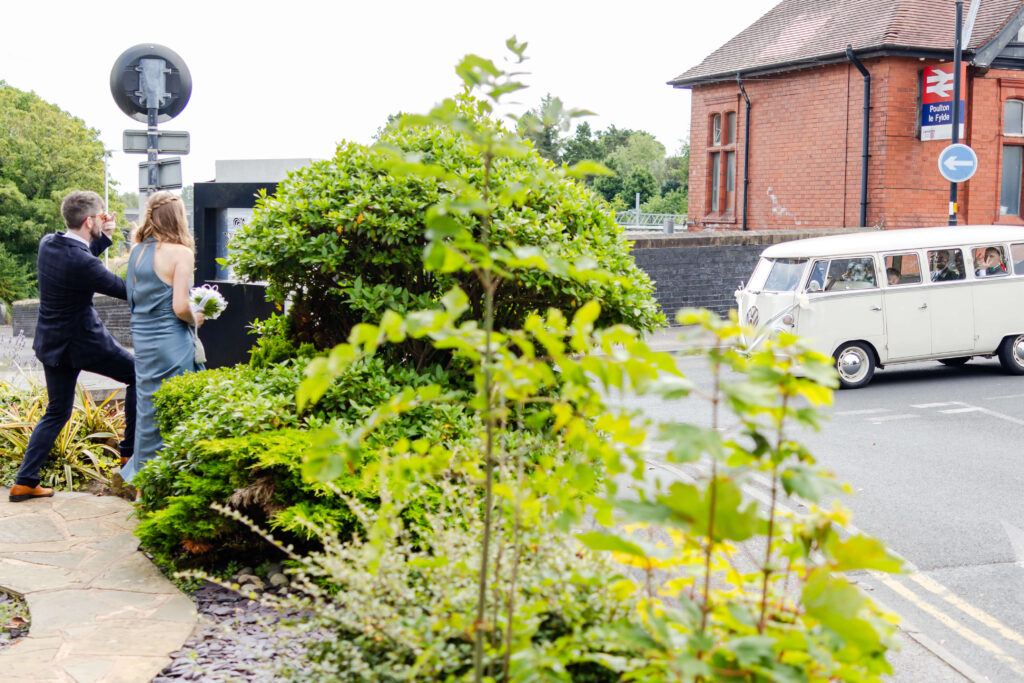A man and woman in formal attire walk past greenery towards a street as a vintage white van drives by near a brick building and a railway station sign. Copyright Somerside Photography Ltd // www.somersidephotography.com