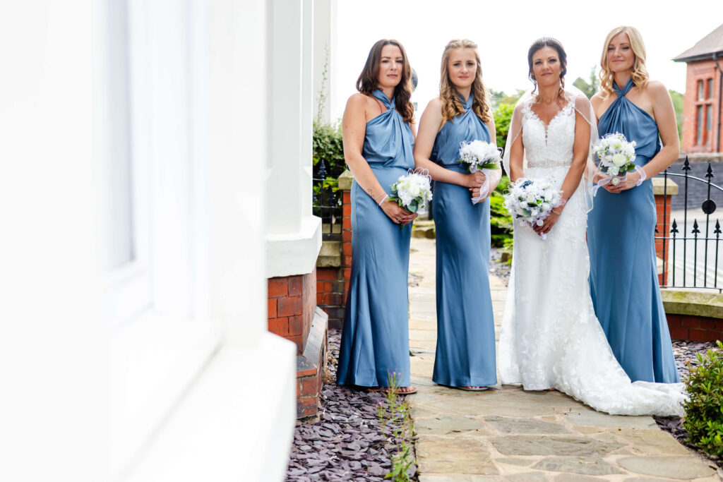 Four women pose outdoors; three wear matching blue dresses holding bouquets, one wears a white bridal gown and holds a bouquet. Copyright Somerside Photography Ltd // www.somersidephotography.com