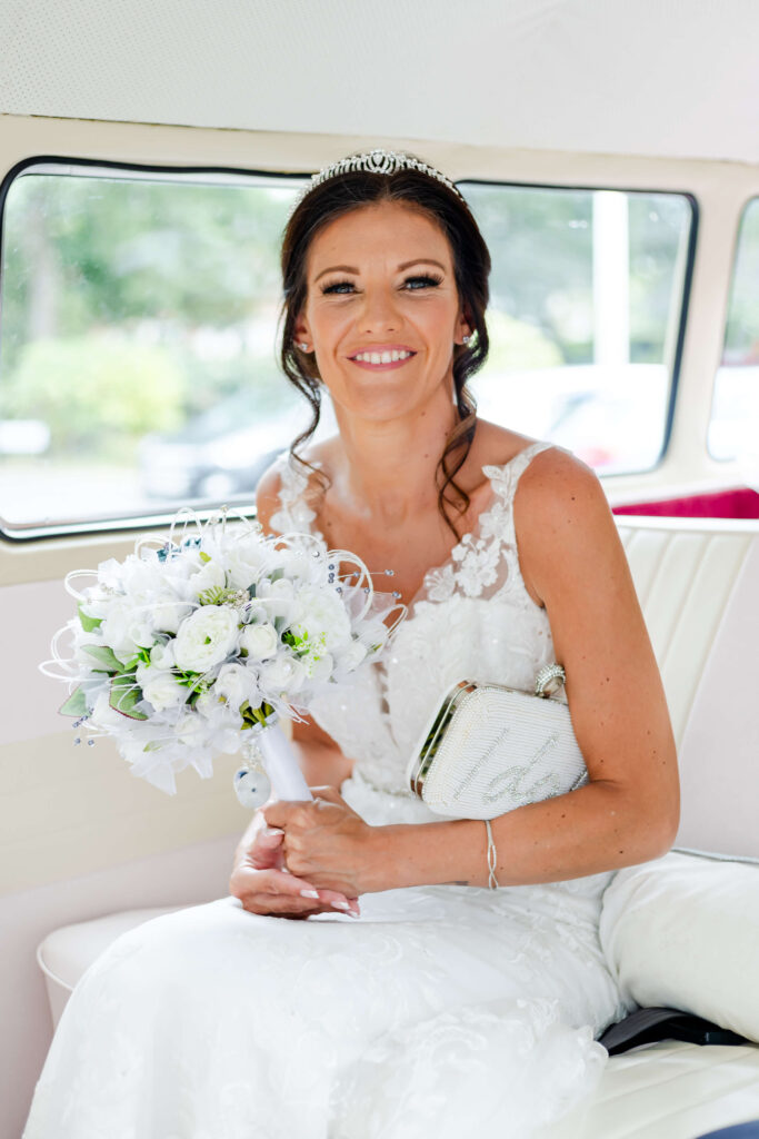 A bride in a white lace dress sits on a bench holding a bouquet of white flowers and a clutch, smiling at the camera. Copyright Somerside Photography Ltd // www.somersidephotography.com