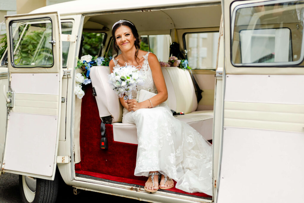 A bride in a white wedding dress sits inside a vintage van decorated with flowers, holding a bouquet and smiling at the camera. Copyright Somerside Photography Ltd // www.somersidephotography.com