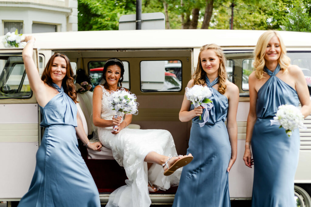 Four women in blue dresses and a bride in a white gown pose cheerfully outside a cream-coloured van, holding bouquets, with greenery and buildings in the background. Copyright Somerside Photography Ltd // www.somersidephotography.com