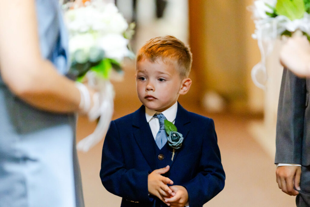 A young boy in a blue suit and tie stands indoors, holding his hands together, surrounded by people holding bunches of flowers. Copyright Somerside Photography Ltd // www.somersidephotography.com