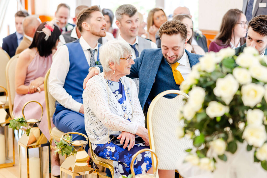 A group of people sit together at a wedding ceremony, with a young man smiling and putting his arm round an elderly woman in the front row. White flowers are visible in the foreground. Copyright Somerside Photography Ltd // www.somersidephotography.com