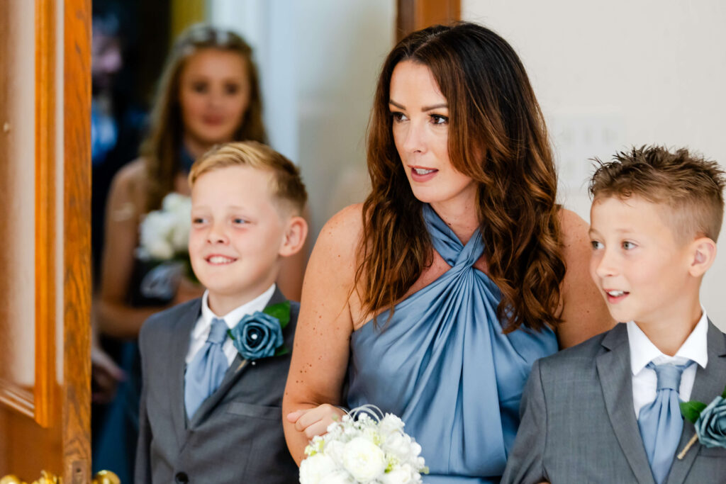 A woman in a blue dress holding a bouquet stands between two boys in grey suits with buttonholes, all looking ahead indoors. Copyright Somerside Photography Ltd // www.somersidephotography.com