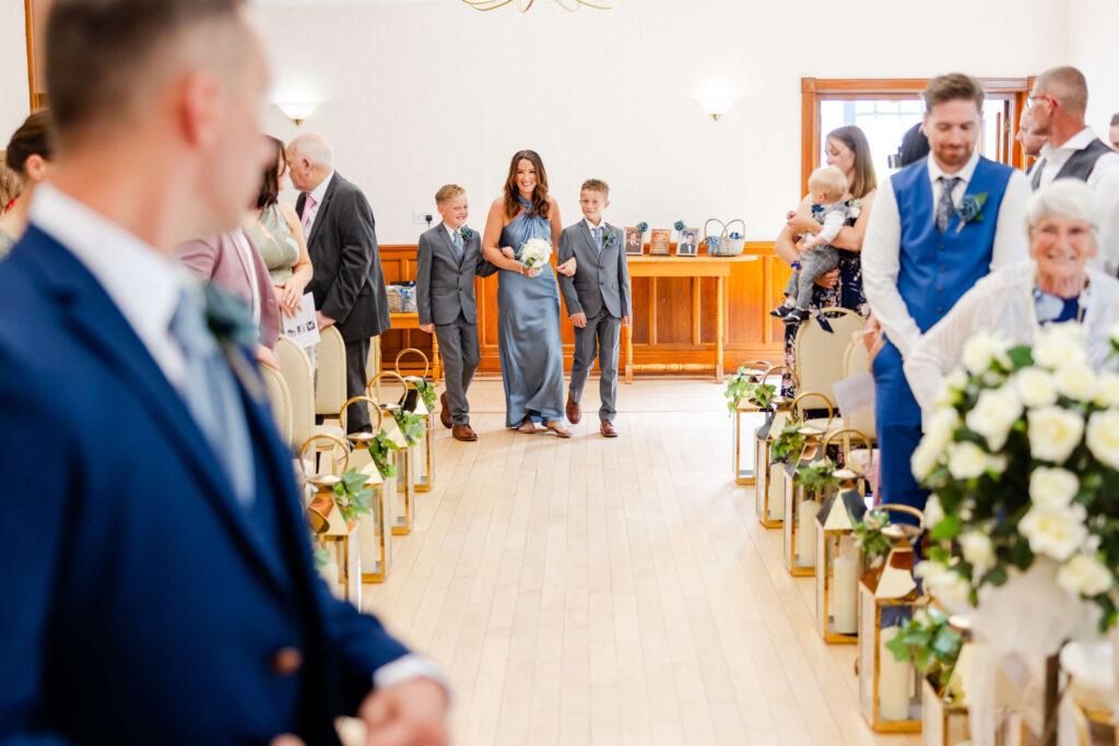 A woman in a blue dress walks down the aisle with two boys in suits at a wedding ceremony, while guests look on. Copyright Somerside Photography Ltd // www.somersidephotography.com