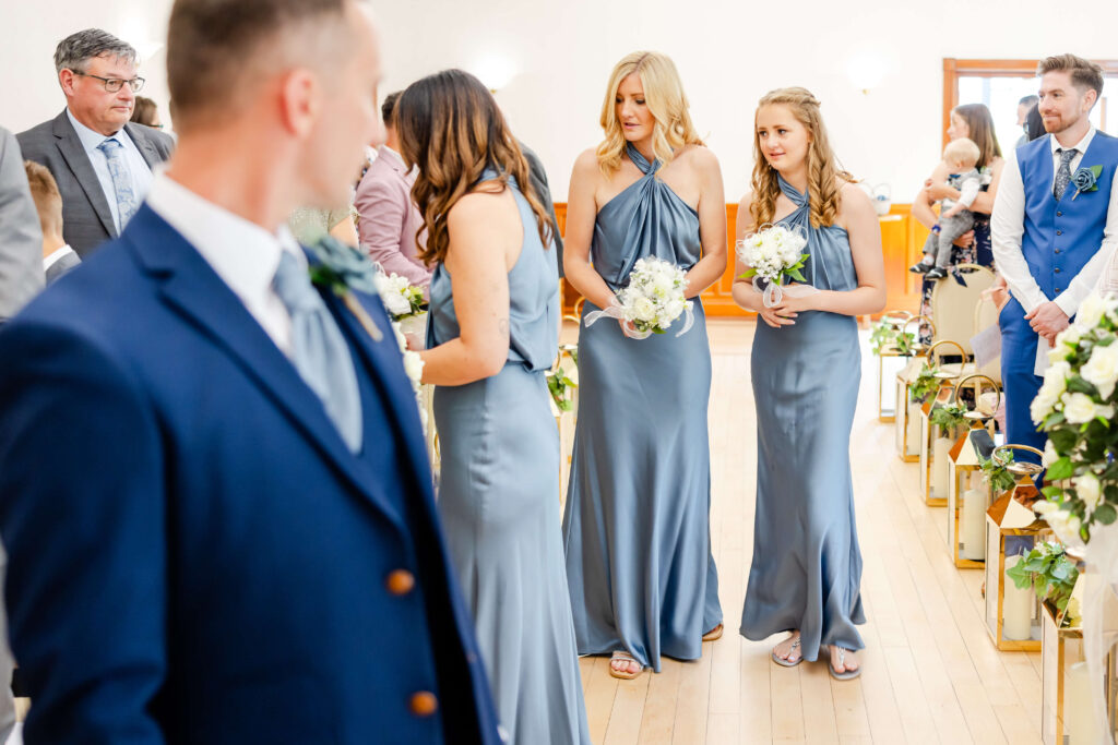 Bridesmaids in blue dresses walk down the aisle holding bouquets, while guests and groomsmen stand and watch in a brightly lit indoor wedding venue. Copyright Somerside Photography Ltd // www.somersidephotography.com