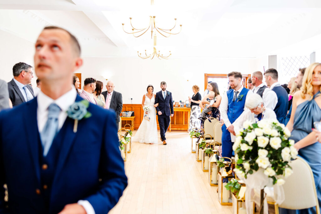 A bride walks down the aisle with a man in a suit, guests seated and standing on either side in a bright, decorated wedding venue. Copyright Somerside Photography Ltd // www.somersidephotography.com