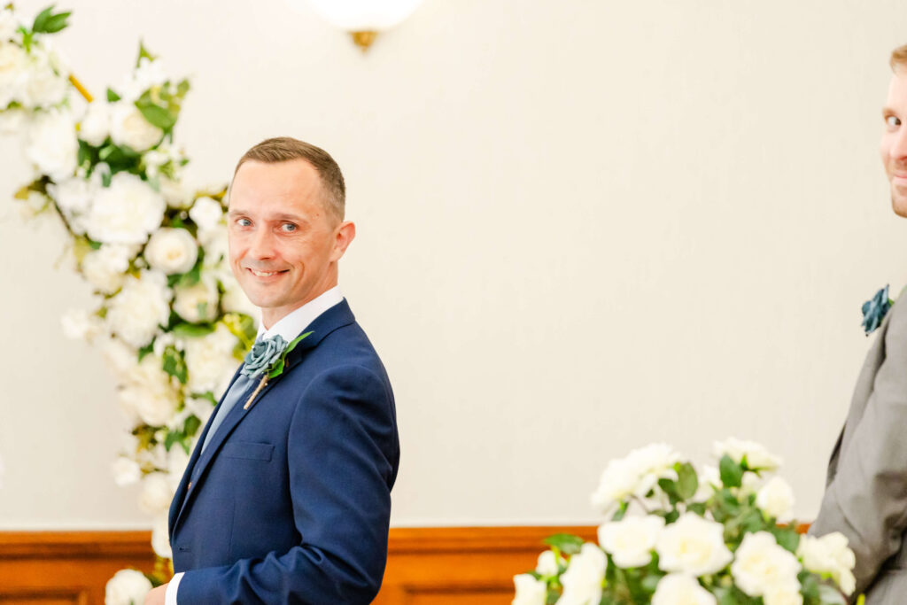A man in a navy suit smiles whilst standing indoors near white floral arrangements. Another person in a grey suit is partially visible beside him. Copyright Somerside Photography Ltd // www.somersidephotography.com