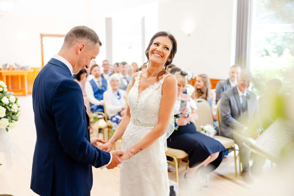 A bride and groom stand holding hands and smiling during their wedding ceremony, with seated guests watching in a bright room. Copyright Somerside Photography Ltd // www.somersidephotography.com